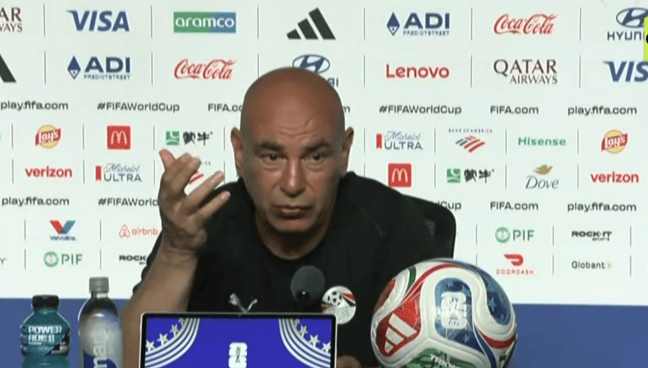 Bald man at a press conference gesturing with his hand, seated behind a table with a soccer ball and laptop screen in front of him.
