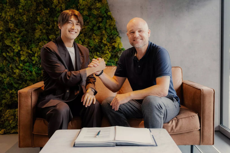 Two men smile and shake hands on a brown sofa in a modern office with a green moss wall behind them and documents on a coffee table in front of them