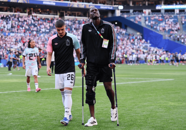 Two soccer players walk across the field after a match; one is on crutches with a knee brace, the other in a black and pink jersey, crowd in the stands behind them.