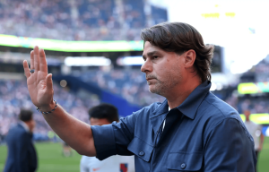 Coach in a blue shirt raises his hand to gesture on a football field with blurred stadium crowds in the background Patiently as fans cheer in the stands.