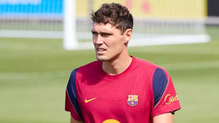 Soccer player in a red FC Barcelona training jersey on a grassy pitch, looking to the side with a goal blurred in the background.