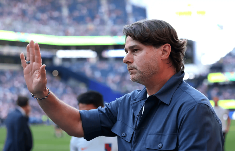Coach in a blue shirt raises his hand to gesture on a football field with blurred stadium crowds in the background Patiently as fans cheer in the stands.