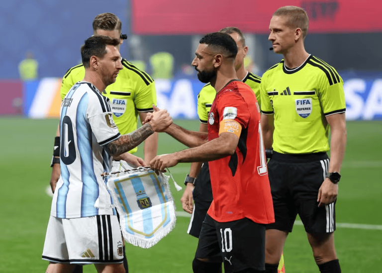 Two soccer players shake hands on the field before kickoff, one wearing a light blue-and-white striped jersey and the other in red, with FIFA referees in bright yellow uniforms nearby.