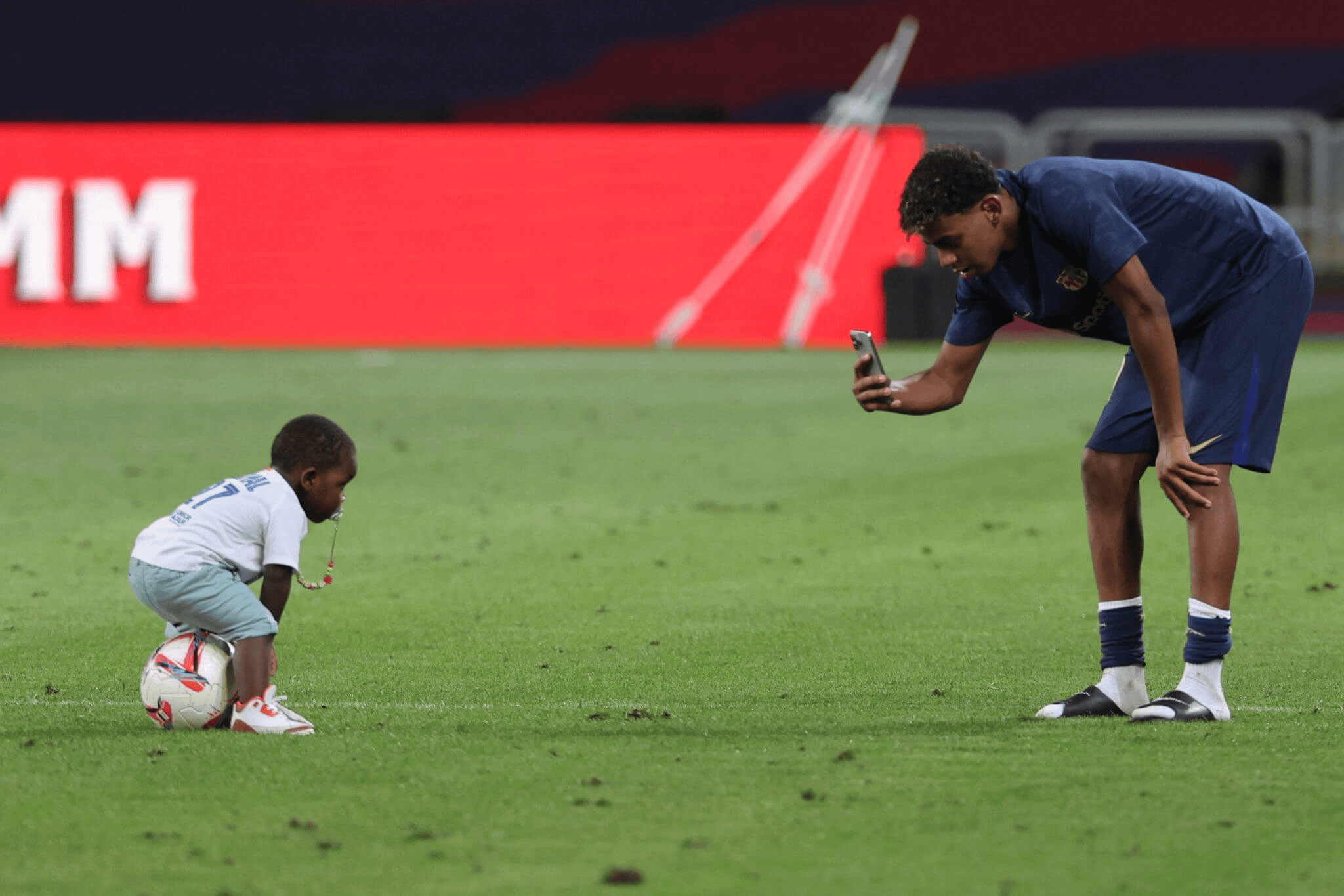 A small child kneels on a soccer field with a ball while an adult in a blue uniform crouches to take a photo with a smartphone nearby.
