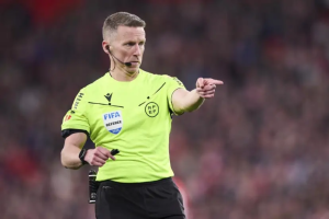 Soccer referee in a neon yellow shirt points toward the field during a match with a headset mic on.