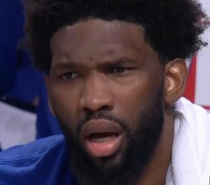 Close-up of a Black man with a beard and short curly hair, mouth open in a surprised or shocked expression, wearing a blue jersey.