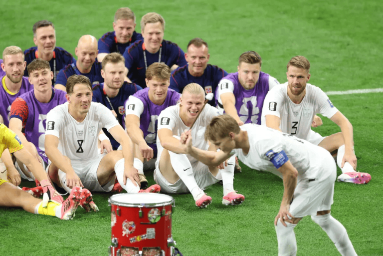 Group of soccer players in white and purple kits posing on a green field after a match, with a red drum in the foreground.
