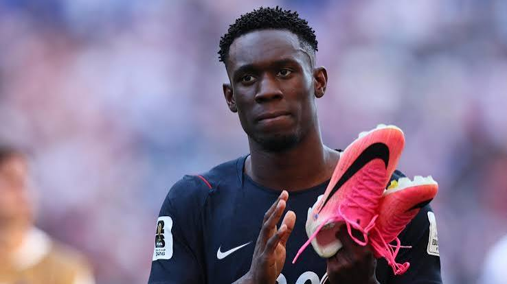 Soccer player in a dark jersey claps while holding bright pink cleats on the field after a match.
