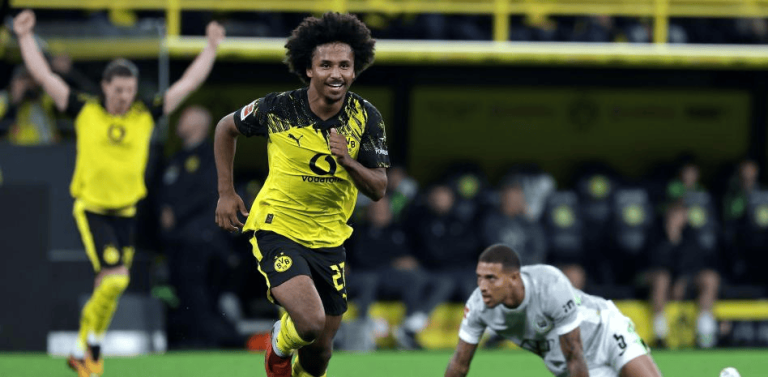Soccer scene with a Borussia Dortmund player in yellow and black sprinting, while an opponent in white lies on the ground nearby on the field.