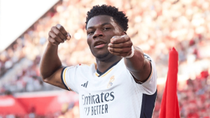 Real Madrid footballer in a white kit celebrating on the field, pointing toward the camera with both hands as the crowded stadium cheers.