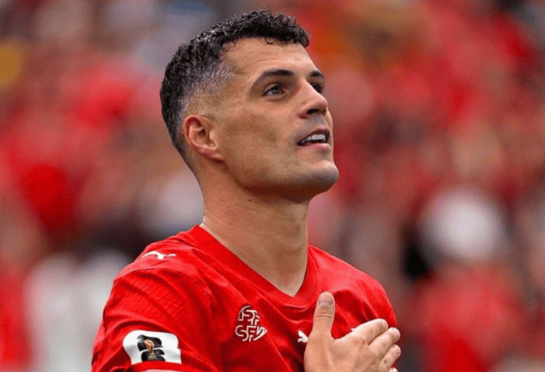 Soccer player in a red jersey looks upward with his right hand over his heart on a crowded stadium background.