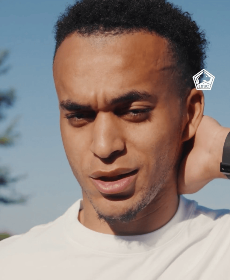 Close-up of a man with short curly hair in a white shirt, outdoors on a sunny day, hand resting on his neck, LOSC logo visible near his ear.