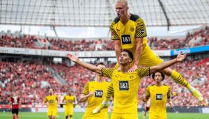 Borussia Dortmund players celebrate a goal in yellow kits on the pitch, with one player lifted on another's shoulders and a packed stadium behind them.