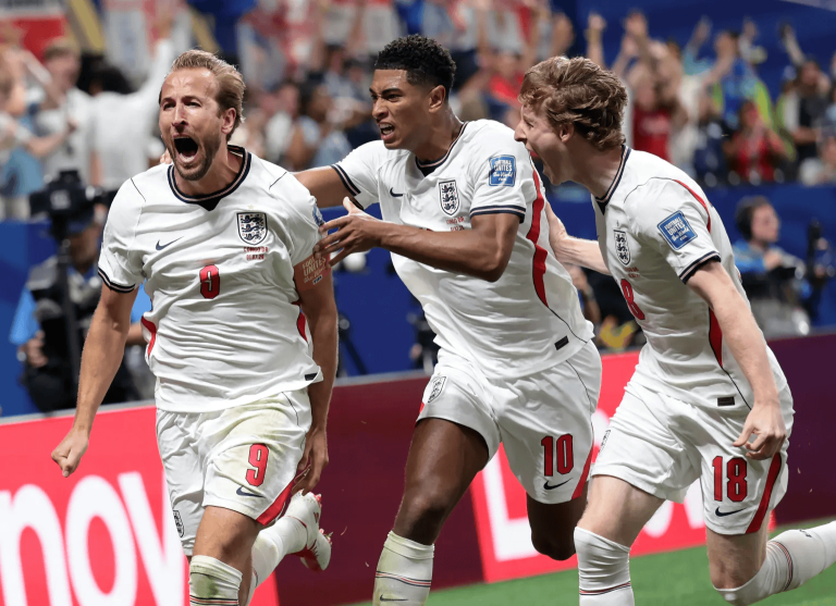 Three England soccer players celebrate a goal, running together on the pitch with arms around each other.