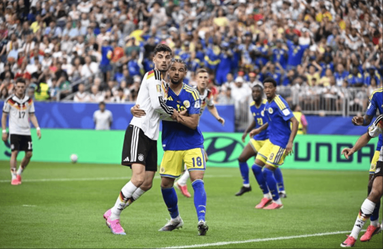 Two soccer players, one in a white Germany kit and one in blue Colombia kit, grapple for the ball on a green pitch with a crowded stadium behind them.