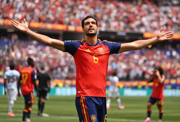 Soccer player wearing a red Spain jersey with arms outstretched in celebration on a green field, full stadium cheering in the background.