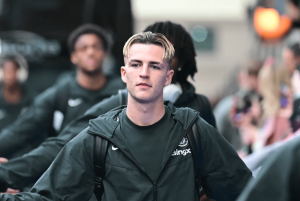 Young man with blond hair in a dark green windbreaker walking through a crowded street.