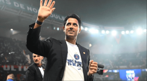 Man in a blazer waves to the crowd on a stadium stage, wearing a white T-shirt with text.