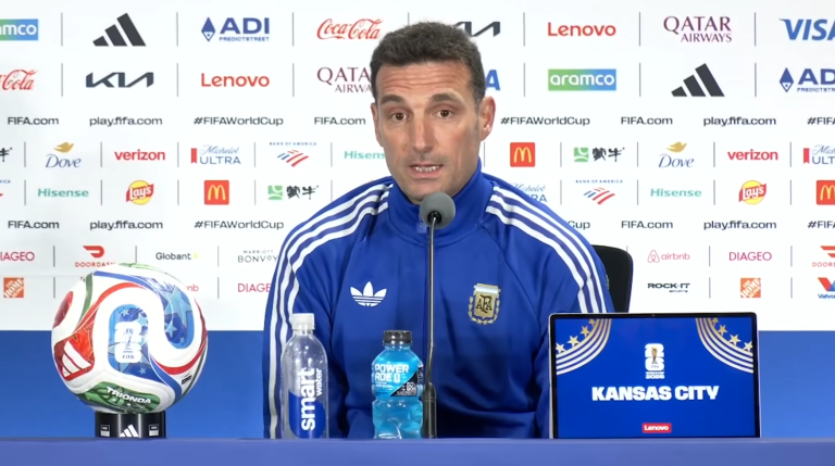 Argentine coach speaks at a FIFA World Cup press conference, with a colorful soccer ball on a stand and a laptop on the table.