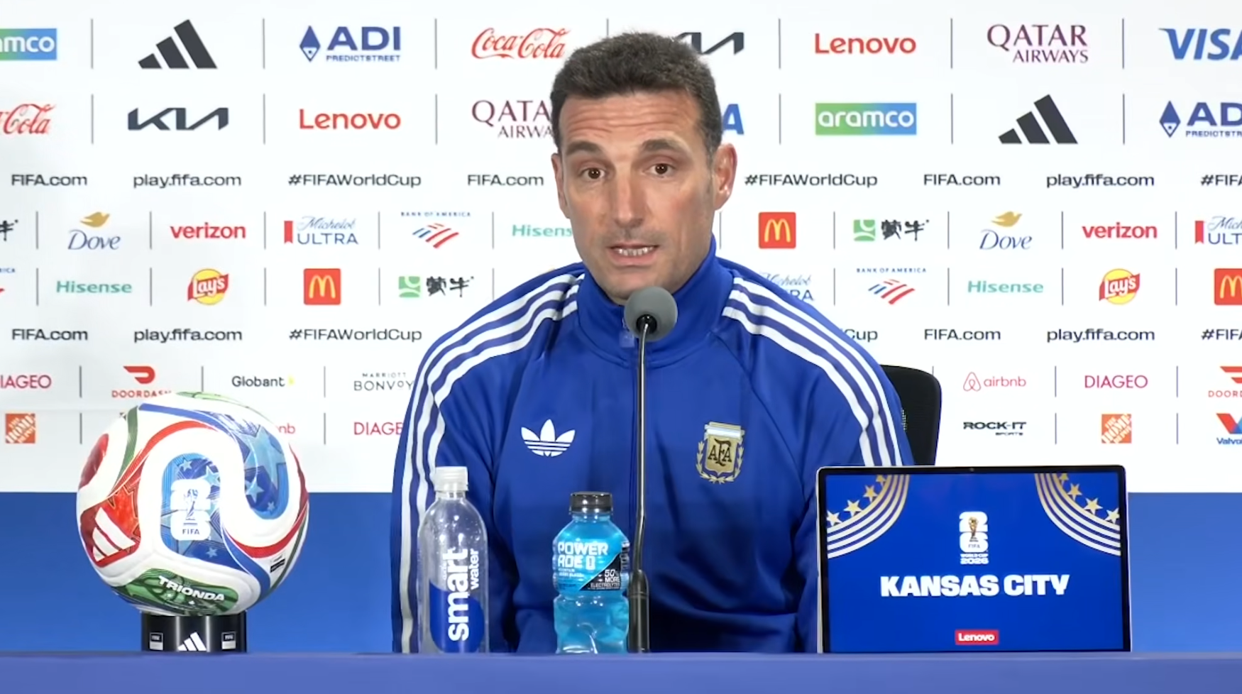 Argentine coach speaks at a FIFA World Cup press conference, with a colorful soccer ball on a stand and a laptop on the table.