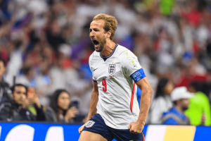 England footballer celebrates a goal, mouth open and fist clenched, wearing white jersey with number 9 and captain's armband, crowd in the background