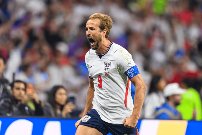 England footballer celebrates a goal, mouth open and fist clenched, wearing white jersey with number 9 and captain's armband, crowd in the background