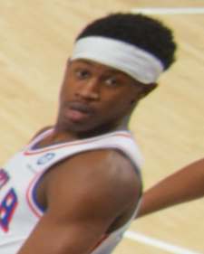 Basketball player wearing a white headband and jersey on a court, looking toward the camera.