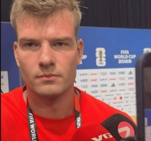 Close-up of a young man in a red shirt being interviewed, microphone to the right, FIFA World Cup 2026 backdrop in blue nearby.