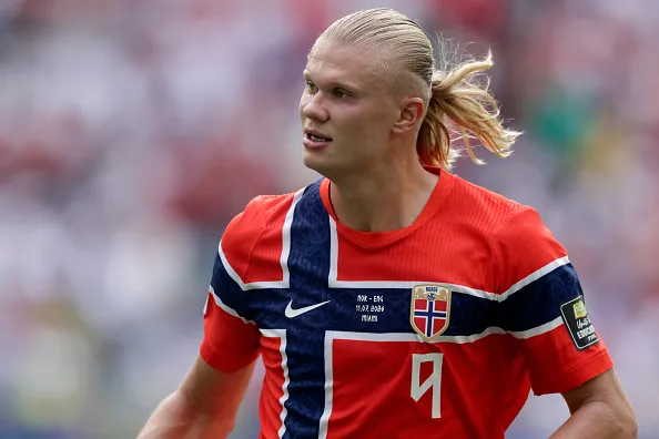 Male soccer player in a red jersey with a Norwegian flag crest, sprinting on a blurred stadium field.