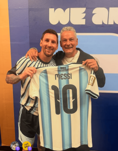 Two men pose together, holding up an Argentina national team jersey with the name and number 10, in front of a blue and orange backdrop labeled 'WE ARE'.