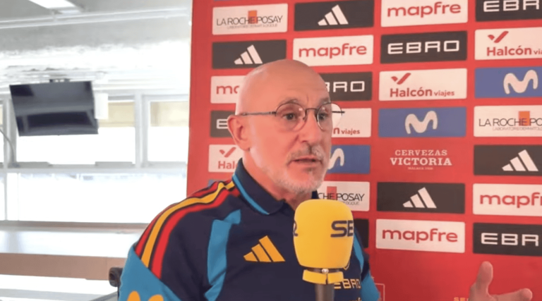 Man with glasses being interviewed in front of a red sponsor backdrop, speaking into a yellow microphone.
