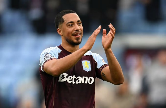 Aston Villa footballer in maroon and light blue kit clapping with hands raised on a field.
