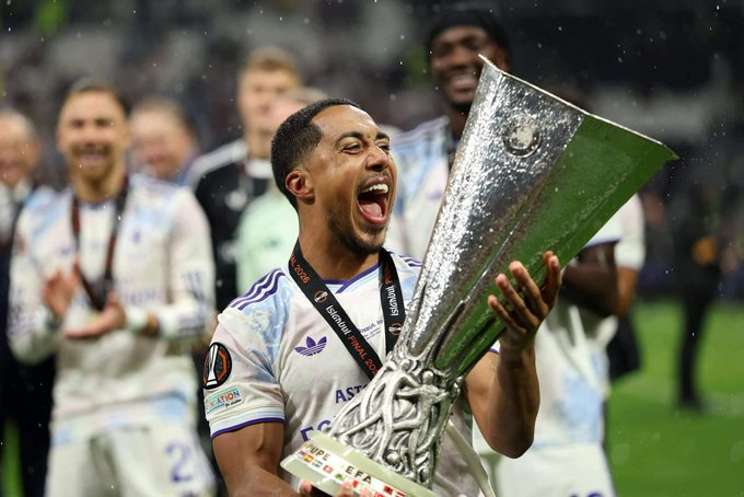 Soccer player in white jersey lifts a large silver trophy in celebration on the field, teammates in the background.