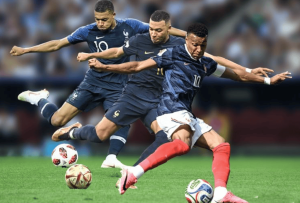 Three France soccer players battle for the ball on a green field; the foreground player in a blue jersey with red socks kicks the ball as teammates close in.