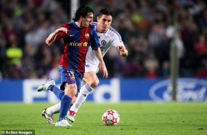 Barcelona player in red-and-blue kit dribbles the ball while a Chelsea defender in white pursues during a match on a green pitch.
