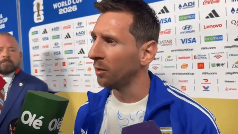 Male athlete in a blue jacket giving an interview, microphones held up in front of a sponsor wall.