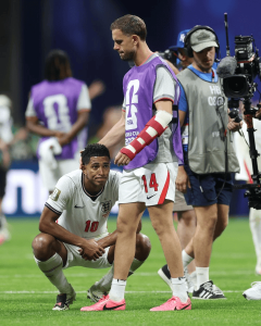Two England footballers on a green pitch during training/entertainment event; one kneels in white kit (#10) while another stands with a purple bib (#14) as a cameraman films in the background.