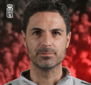 Close-up portrait of a man with short dark hair and light stubble, wearing a light gray zip-up, with a blurred red background and a crest logo in the top-left.