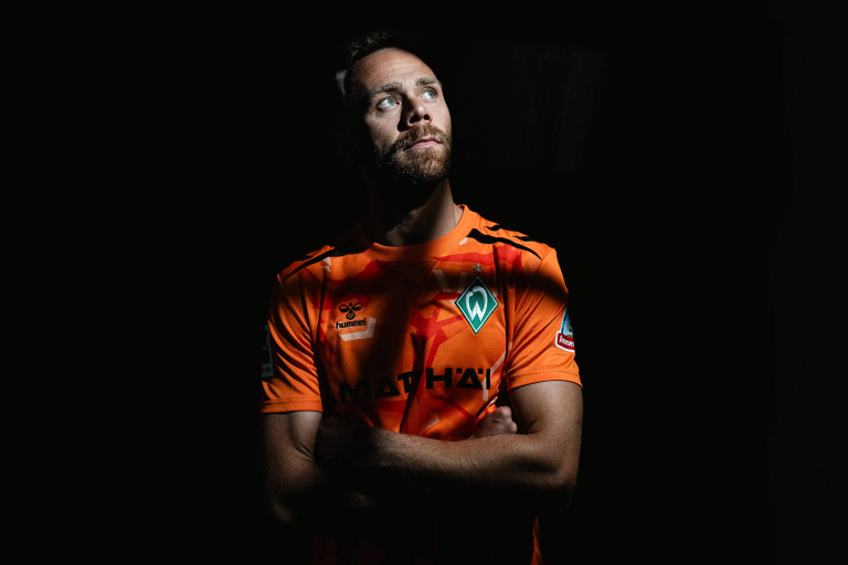 Football player in an orange Werder Bremen jersey, arms crossed, looking upward against a dark background.