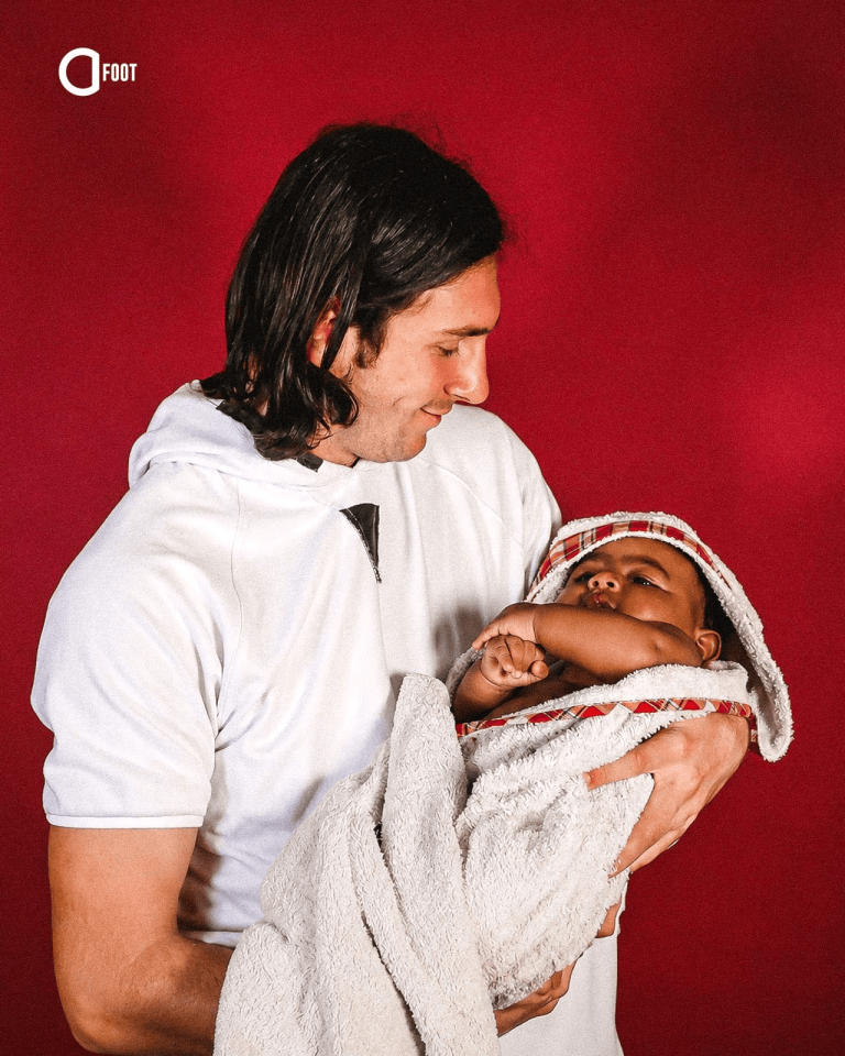 Man in a white shirt cradling a baby wrapped in a white towel against a red backdrop, looking down with a smile.
