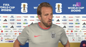 England footballer in a gray jersey speaking at a FIFA World Cup press conference with sponsor logos in the background.