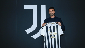 Man in a dark blazer holds up a black-and-white Juventus jersey with Jeep logo against a dark wall.