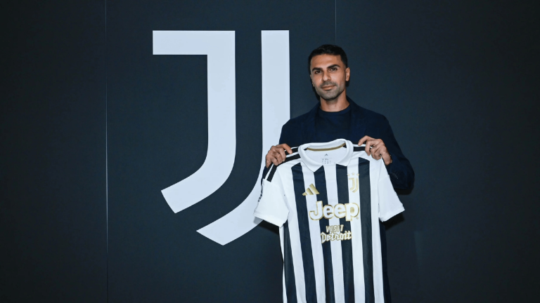 Man in a dark blazer holds up a black-and-white Juventus jersey with Jeep logo against a dark wall.
