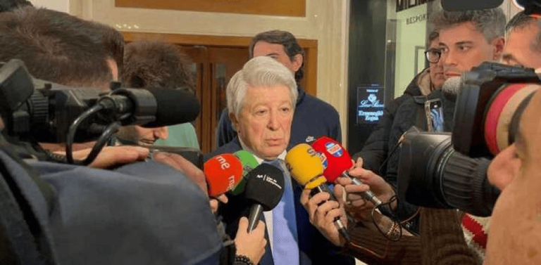 Older man in a suit speaks to the press as reporters hold colorful microphones and cameras surround him.