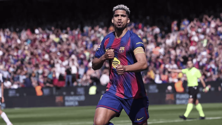Soccer player in Barcelona jersey celebrating a goal on the pitch with a cheering crowd behind him.